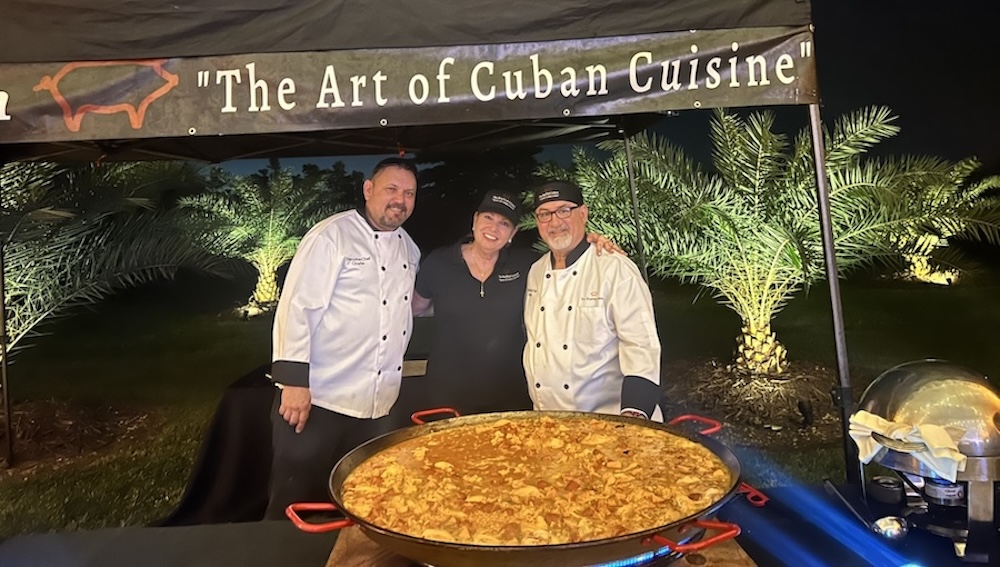Chefs standing behind a large pot of Cuban food and under a banner that says, "The Art of Cuban Cuisine"