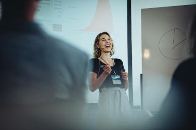 Presenter speaking in front of a projection screen, with audience members out of focus