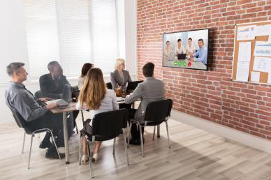 Virtual conference with team members in a room sitting around a table looking at a monitor with a team on the screen