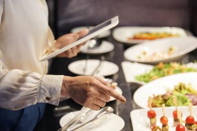 Woman dressed business casual, holding a tablet and pointing to plates of food