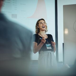 Presenter speaking in front of a projection screen, with audience members out of focus