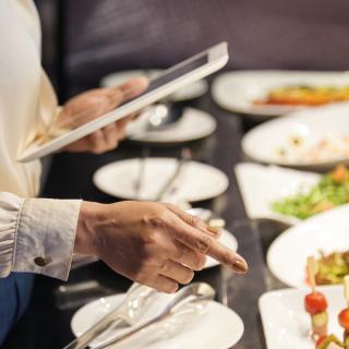 Woman dressed business casual, holding a tablet and pointing to plates of food