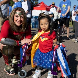 Carolyn Strauss next to a young girl, both dressed up as Wonder Woman. In front of a crowd of people walking
