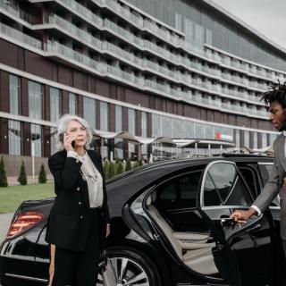 Woman in a suit, on the phone, with a chauffeur opening the door of the limo for her